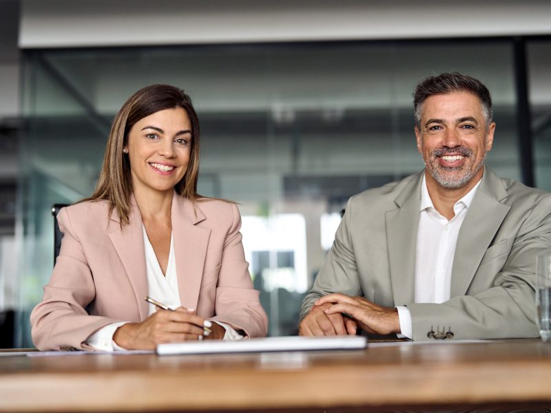 Smiling Latin mature business man and woman sitting at table in office, portrait. Two happy confident professional mid aged corporate executive leaders company managers wearing suits looking at camera